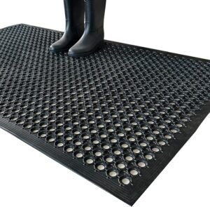Black Uniboho rubber floor mat with circular drainage holes and person in rubber boots standing on anti-slip surface